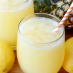 A glass of pineapple lemonade with ice, garnished with a pineapple wedge and lemon slice, against a bright outdoor background.