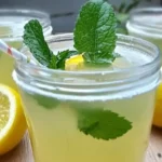 Glass of homemade lemon balm drink with mint leaves and lemon slices, placed on a wooden table with fresh lemons.