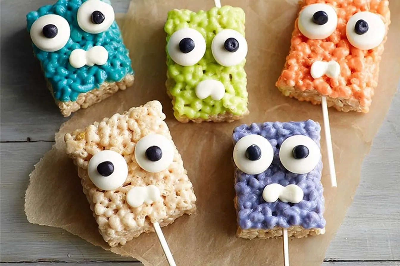 Five colorful Rice Krispie Treat Monsters on sticks, decorated with candy eyes and mouths, arranged on brown parchment paper.