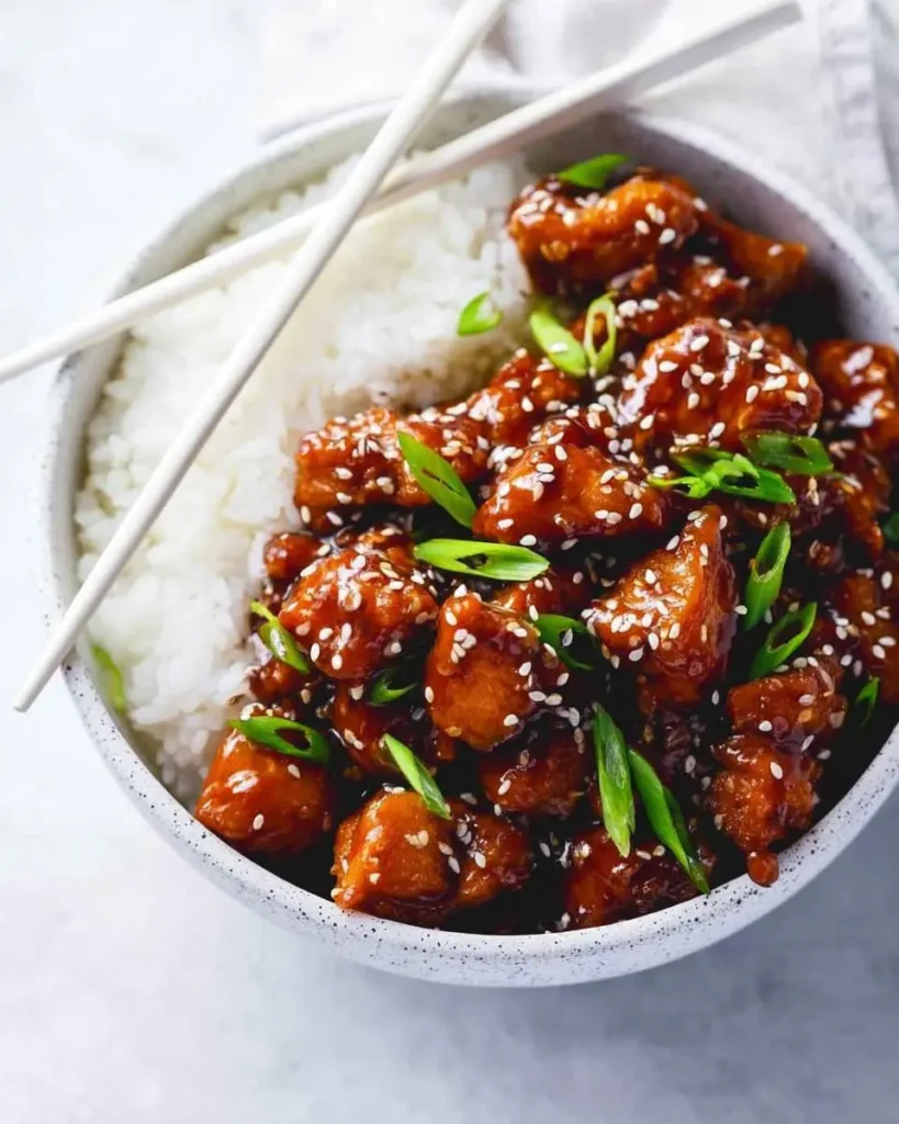 A close-up of a white speckled bowl filled with crispy sesame chicken and white rice, garnished with green onions and chopsticks.