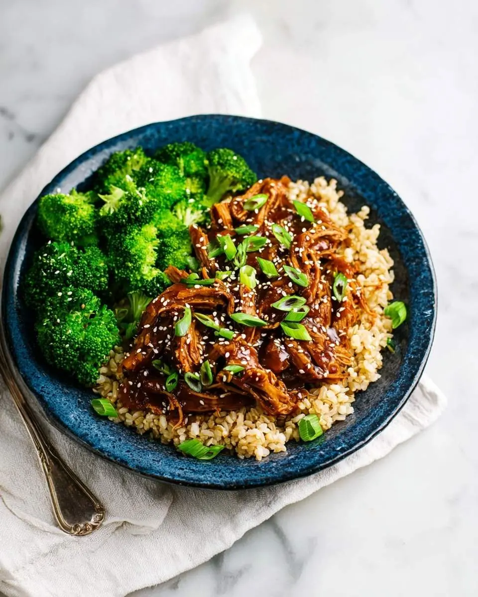 A blue bowl holds a delicious serving of slow cooker honey garlic chicken with brown rice and steamed broccoli, garnished with green onions and sesame seeds.