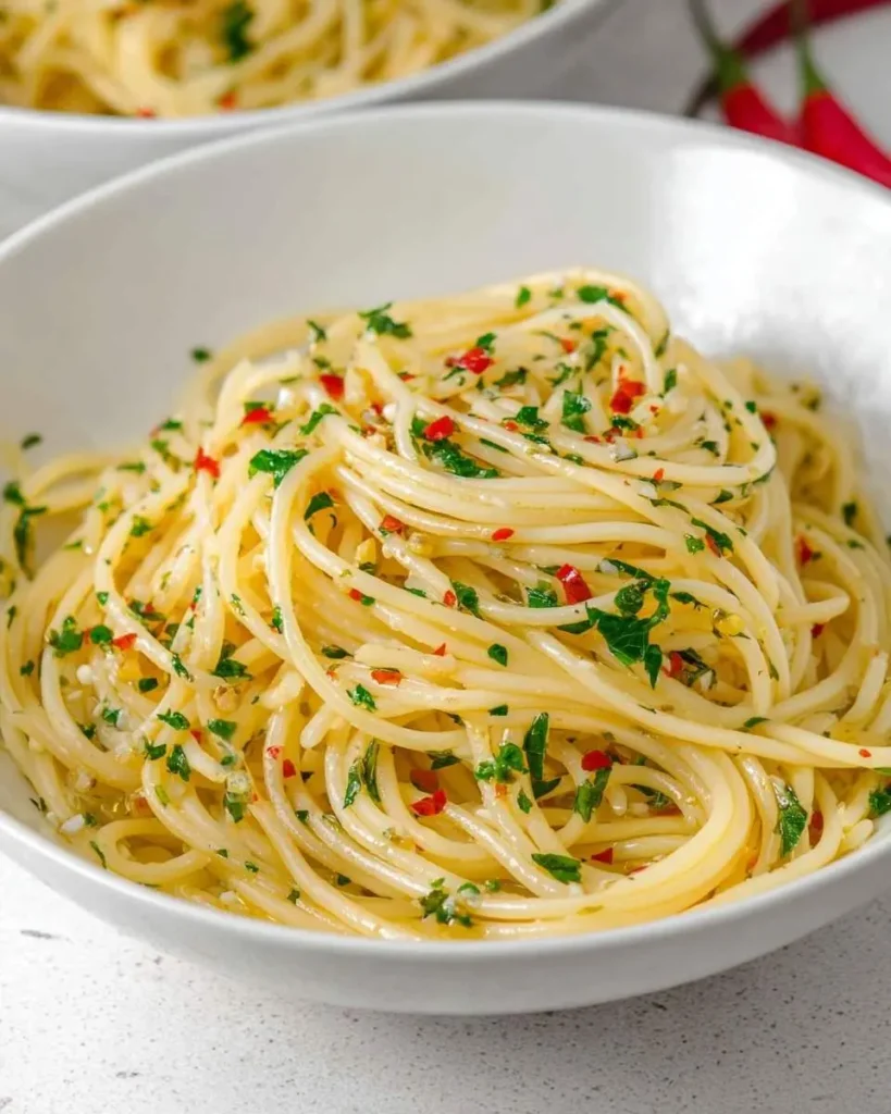 Close-up of a white bowl filled with glistening Spaghetti Aglio e Olio, garnished with fresh parsley and red chili flakes.