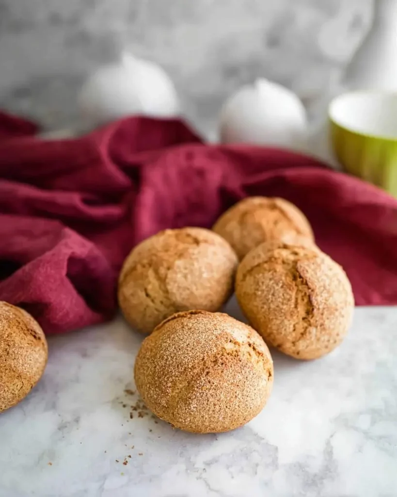 A close-up shows five golden-brown Almond Flour Rolls on a marble countertop next to a red napkin, with blurred kitchenware.