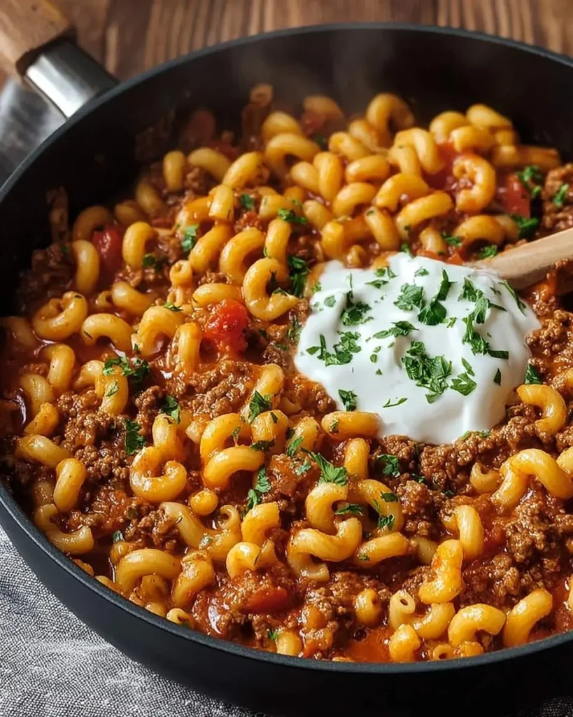 A steaming skillet of hearty Beef Goulash with spiral pasta and ground beef, topped with sour cream and fresh parsley.