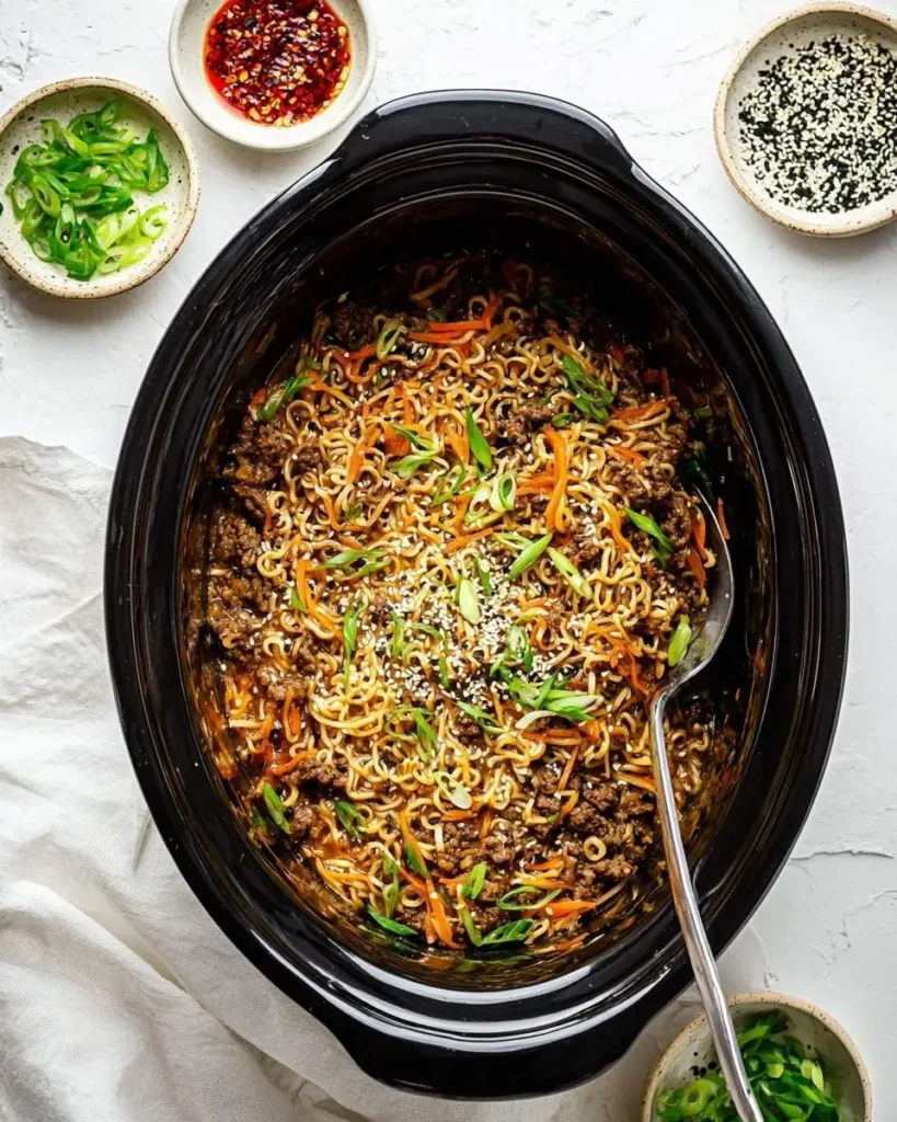 An overhead shot of a large black slow cooker filled with Crockpot Beef Ramen, garnished with green onions and sesame seeds.