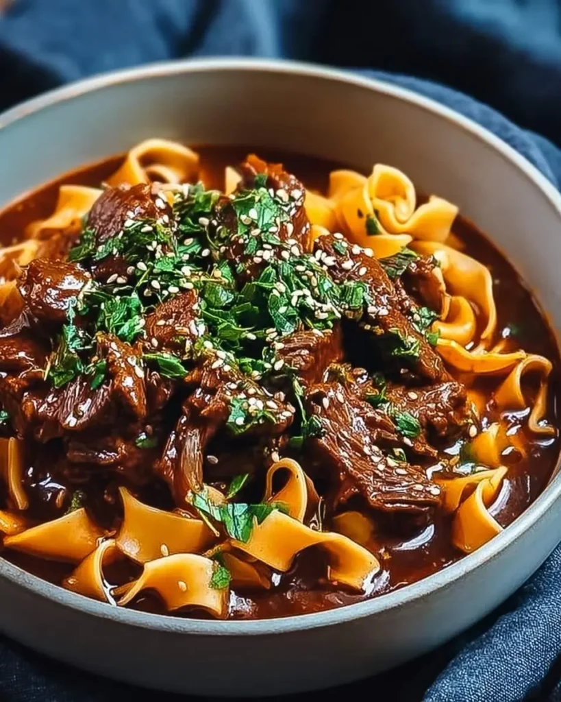 A close-up of a bowl of Korean Beef Noodles, showcasing tender beef, thick noodles, rich dark broth, herbs, and sesame seeds.