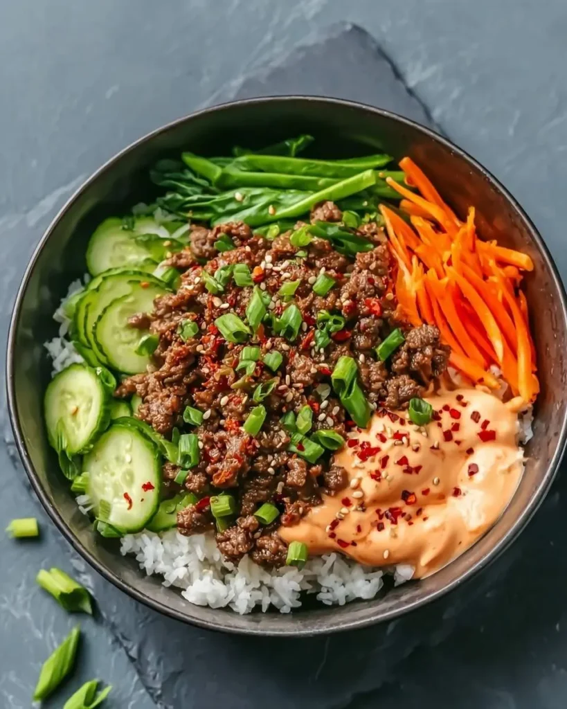 A vibrant Korean Ground Beef Bowl featuring seasoned ground beef, white rice, sliced cucumbers, shredded carrots, green onions, and a creamy sauce.