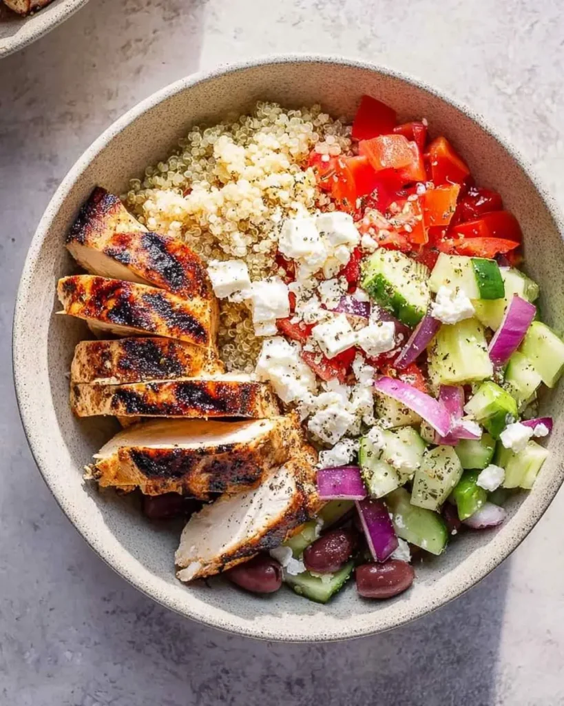 A colorful mediterranean chicken bowl featuring grilled chicken, quinoa, diced tomatoes, cucumbers, feta, and olives.