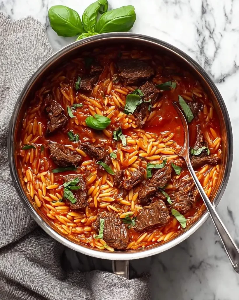 A delicious One Pot Beef Giouvetsi with orzo pasta and tender beef in a rich tomato sauce, garnished with fresh basil, in a silver pot.