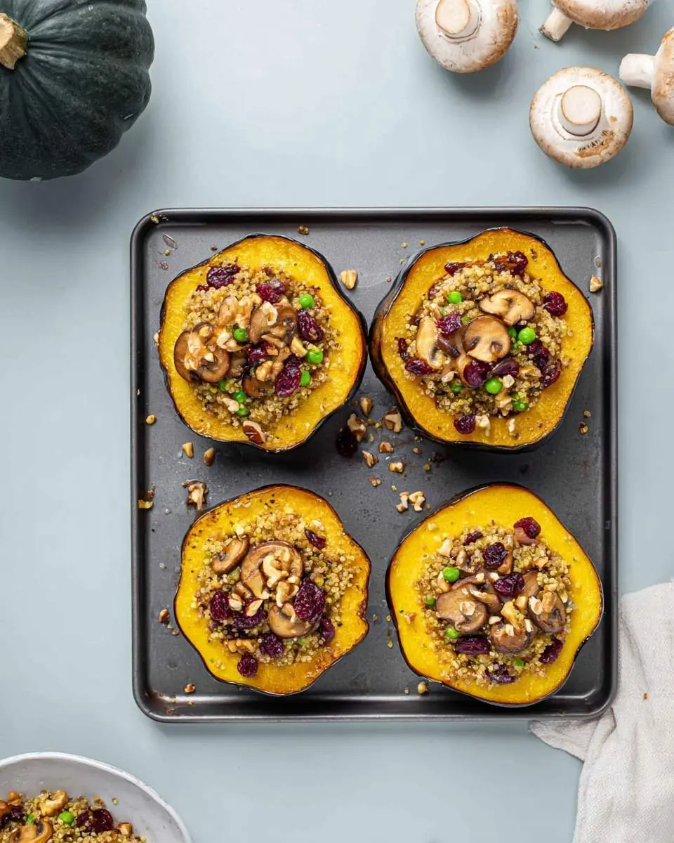 Four halves of Stuffed Acorn Squash on a baking sheet, filled with quinoa, mushrooms, cranberries, peas, and walnuts.