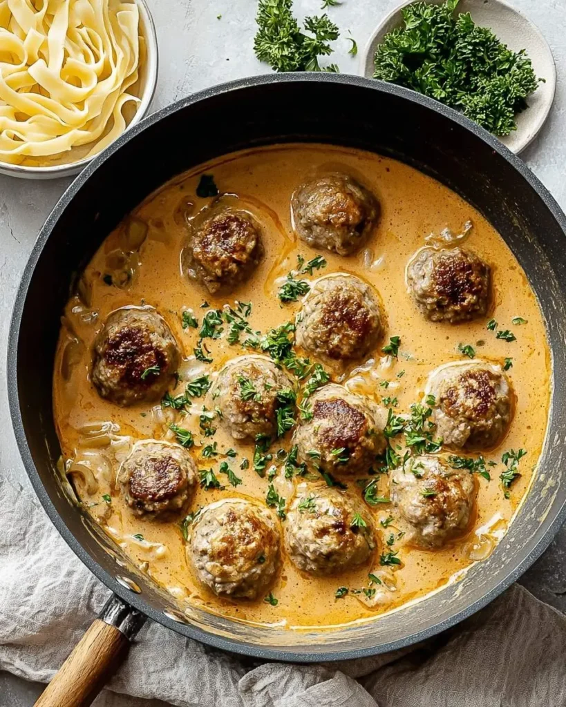 A close-up of a skillet filled with a creamy swedish meatball soup, garnished with fresh parsley and served with a side of pasta.
