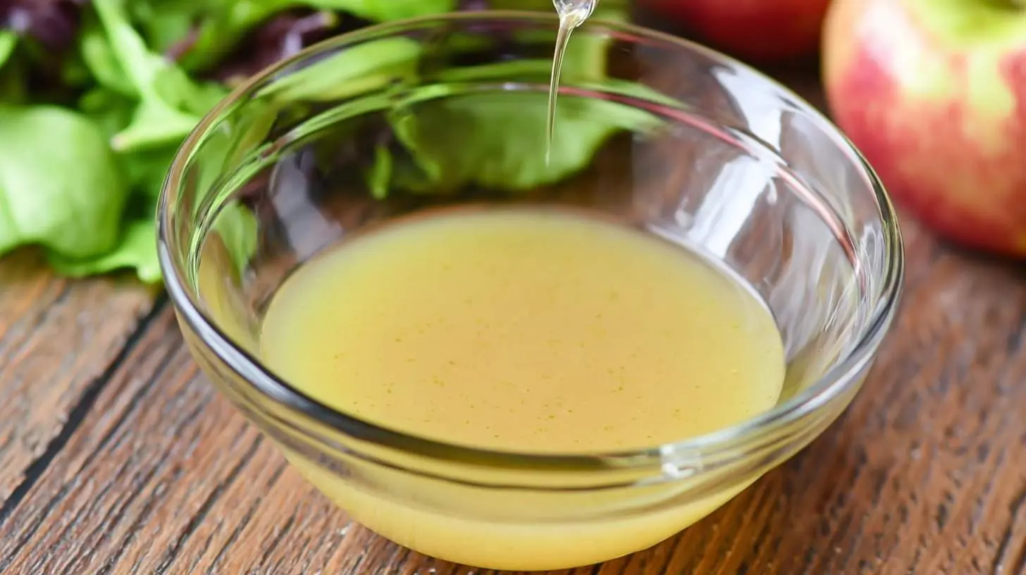 A stream of golden Apple Vinaigrette pours into a clear glass bowl, surrounded by fresh salad greens and apples.