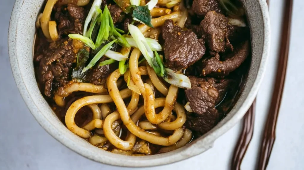 A close-up shot of a bowl filled with savory beef black pepper udon, garnished with fresh green onions and rich sauce.