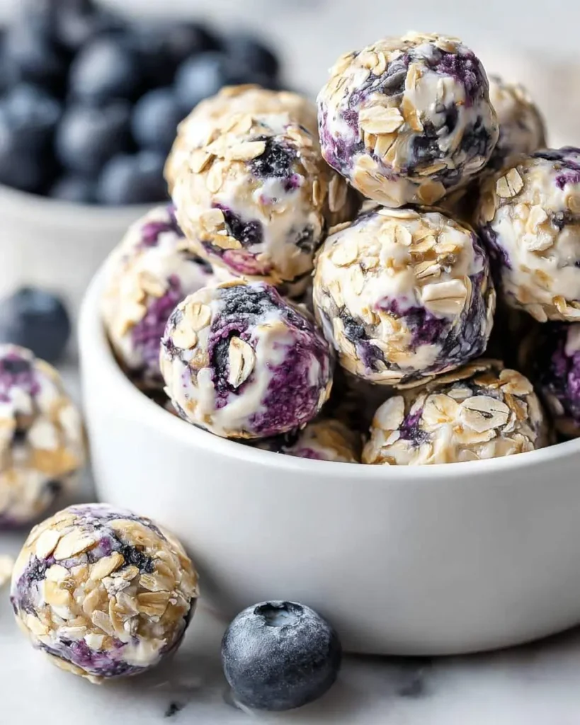 A white bowl overflows with delicious Blueberry Cheesecake Protein Bites, each swirled with purple blueberry and coated in oats, with fresh blueberries nearby.