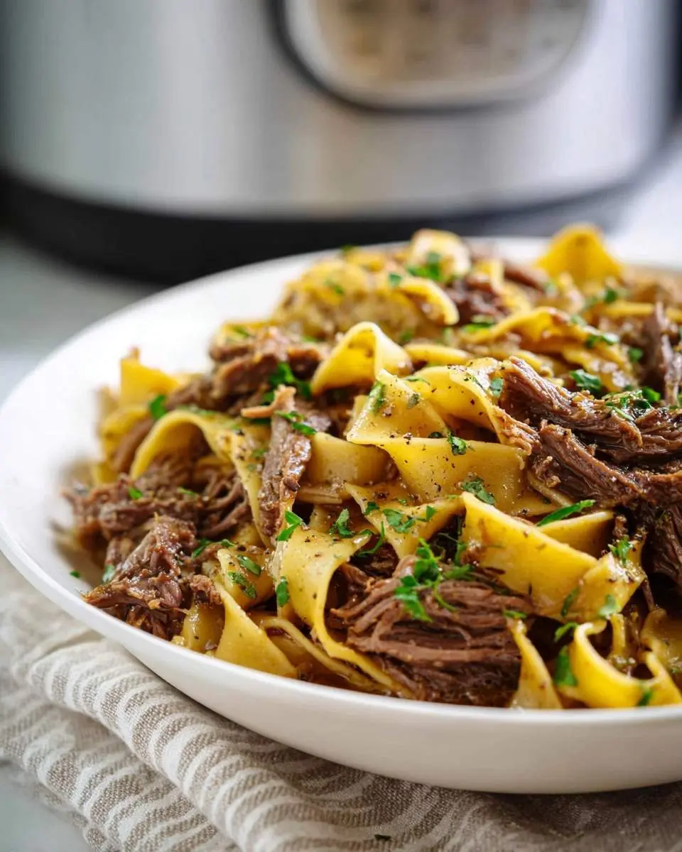A close-up of a white bowl filled with Crock Pot Mississippi Beef Noodles, featuring wide egg noodles tossed with tender shredded beef and fresh parsley, with a blurred Instant Pot in the background.