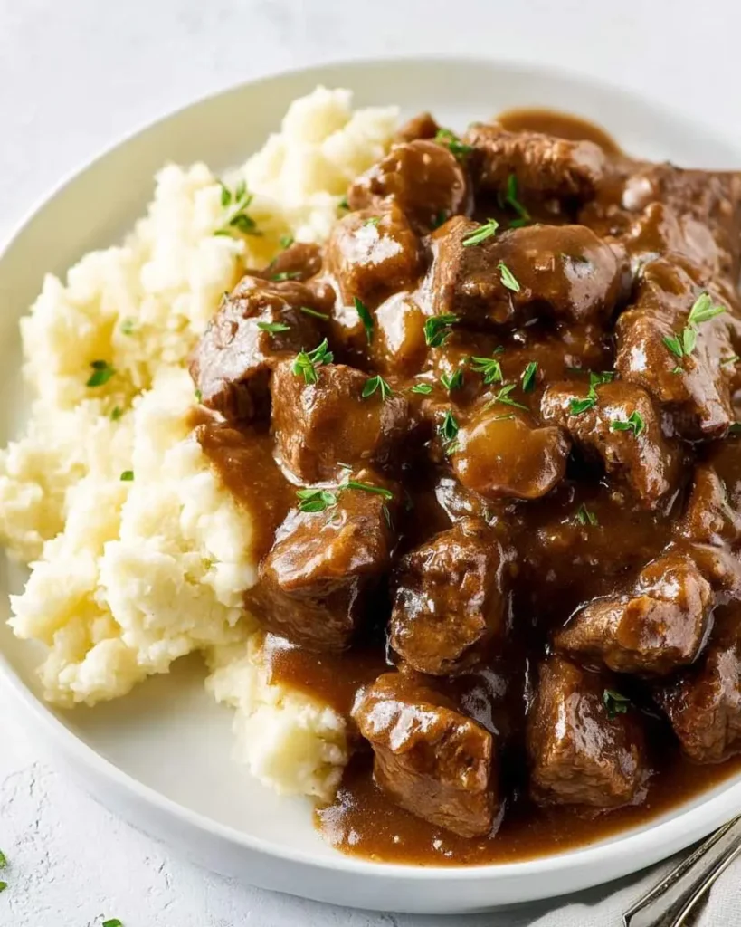 A white plate holds fluffy mashed potatoes next to a generous serving of rich Crockpot Beef Tips With Gravy, garnished with fresh herbs.