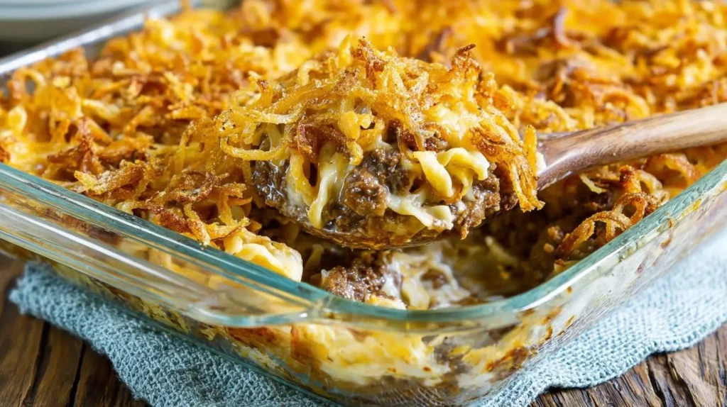 A hearty scoop of french onion beef casserole with cheesy pasta and crispy fried onions being lifted from a glass baking dish.