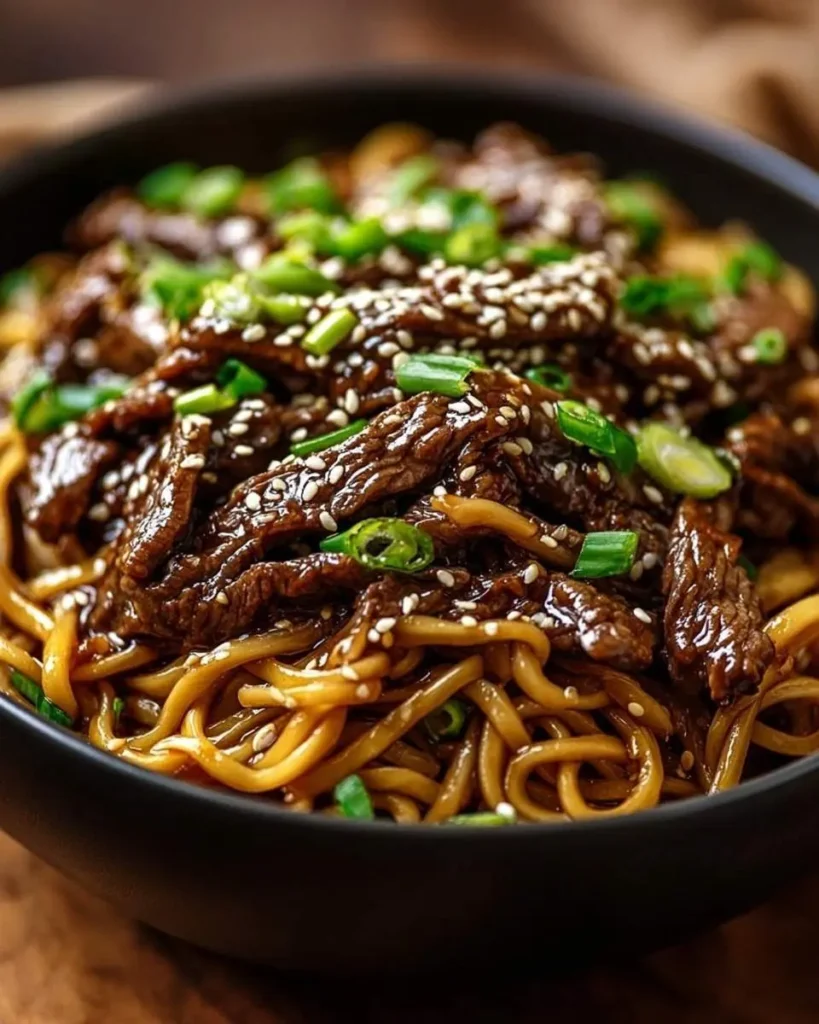 A close-up shot of a black bowl filled with glistening Hoisin Beef Noodles, topped with sesame seeds and chopped green onions.