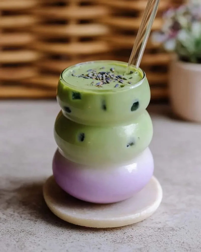 A unique stacked glass with vibrant green Iced Honey Lavender Matcha, topped with lavender buds, and a purple layer at the bottom.