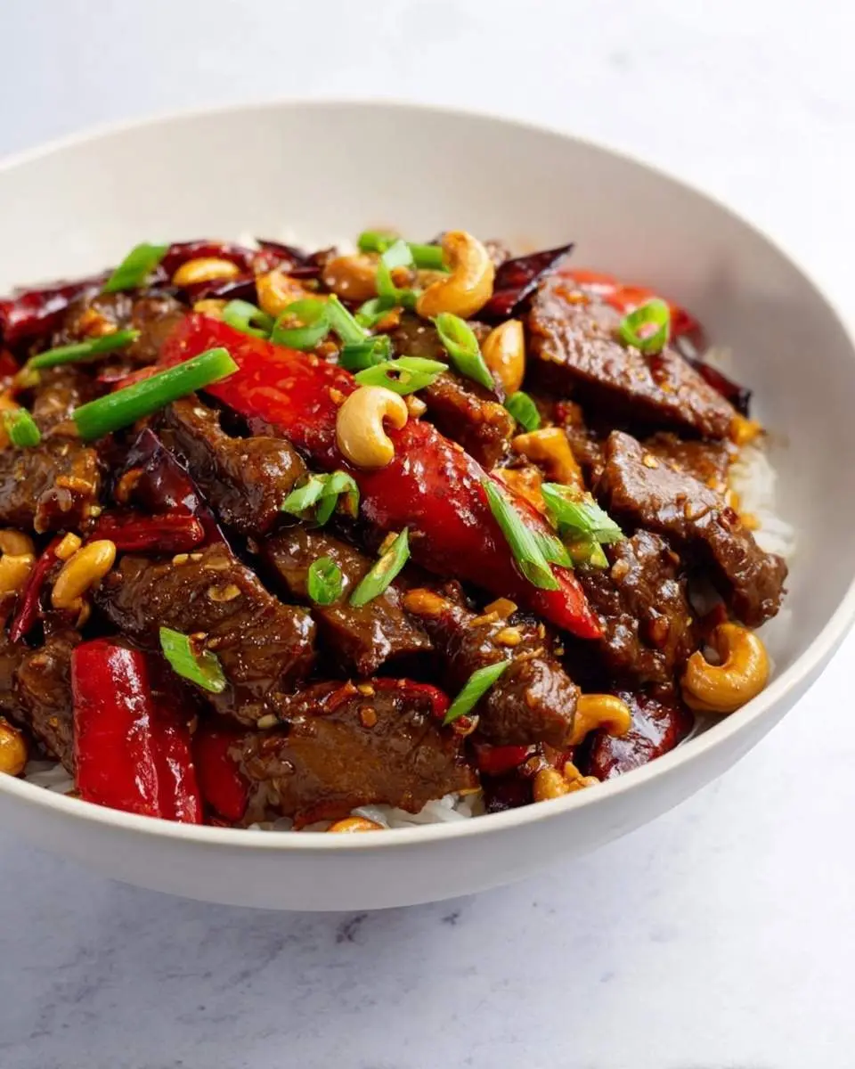 A close-up of a white bowl filled with flavorful Kung Pao Beef, featuring tender beef, red peppers, cashews, and green onions over white rice.