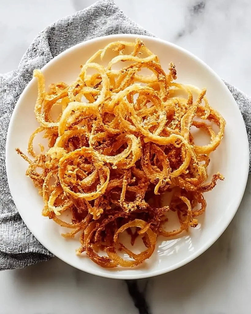 A close-up top-down view of crispy golden-brown onion strings piled high on a white plate, set on a marble surface.