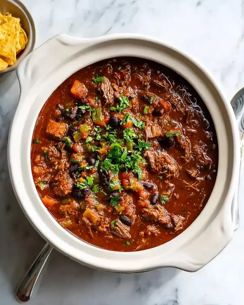 A close-up shot of a white crockpot overflowing with delicious Prime Rib Chili, garnished with fresh herbs, alongside tortilla chips.