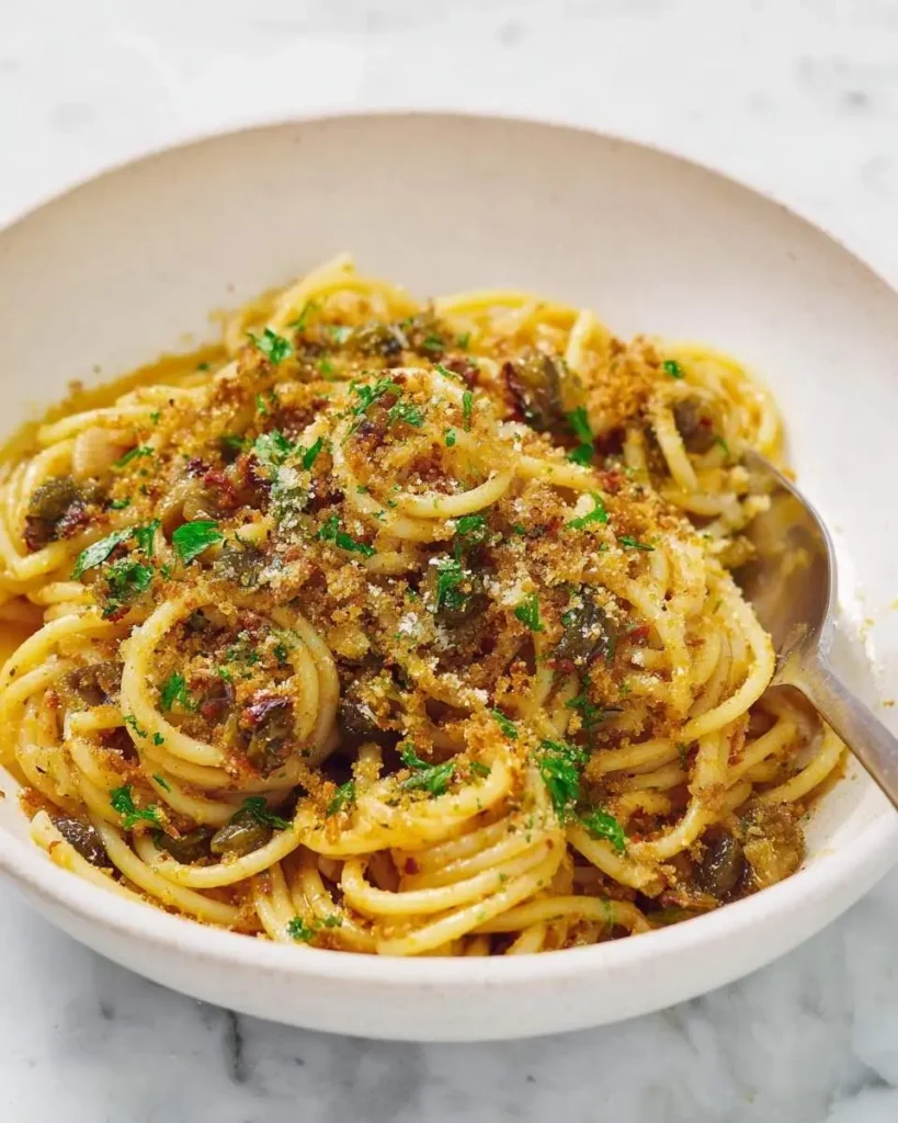 A close-up shot of a white bowl filled with Anchovy Pasta with Breadcrumbs, garnished with parsley and grated cheese.