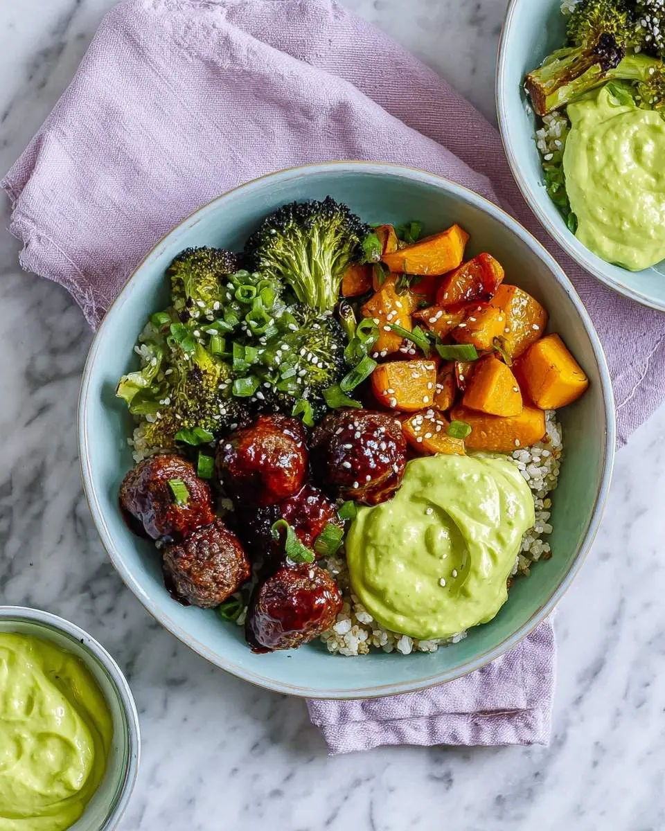 A vibrant Barbecue Meatball Bowl with creamy Avocado Sauce, roasted broccoli, and sweet potatoes on quinoa, garnished with green onions and sesame seeds.