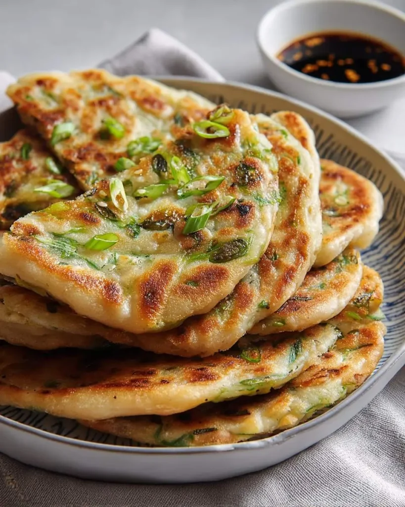 A stack of golden-brown Chinese Scallion Pancakes garnished with fresh green onions and sesame seeds, with a small bowl of dipping sauce in the background.