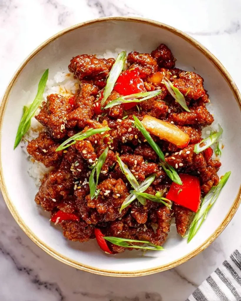 A close-up of a bowl filled with glossy Crispy Beijing Beef, red bell peppers, and green onions over white rice.