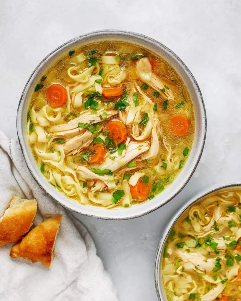 Overhead shot of a delicious bowl of Ginger Garlic Chicken Noodle Soup with wide noodles, shredded chicken, carrots, and fresh herbs, served with crusty bread.