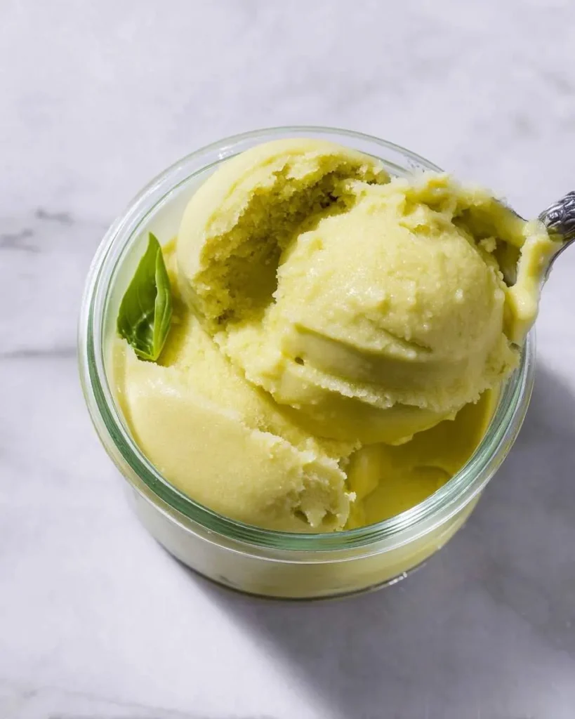 A spoonful of vibrant Ninja Creami Lemon Basil Sorbet being lifted from a clear glass jar, garnished with a fresh basil leaf.