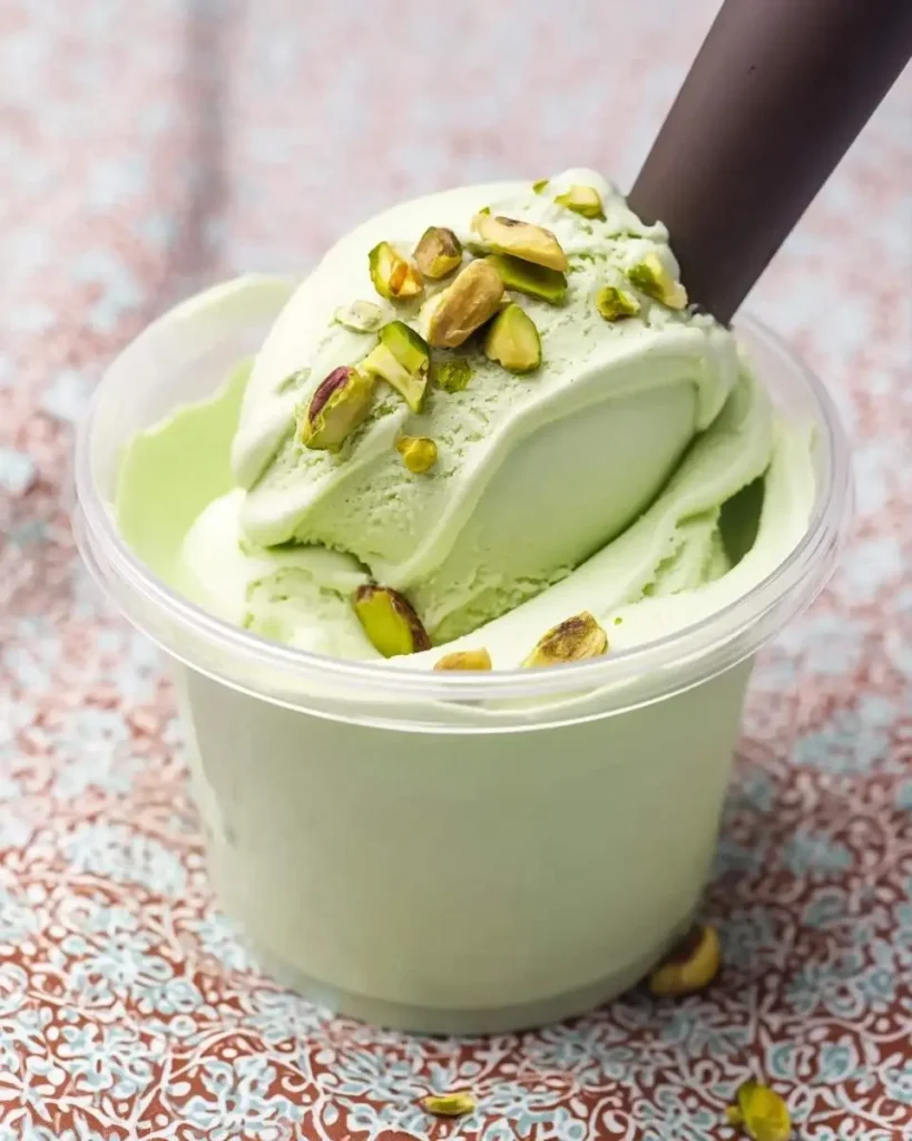 A vibrant scoop of Ninja Creami Pistachio Ice Cream in a clear container, garnished with chopped pistachios, with a dark utensil.