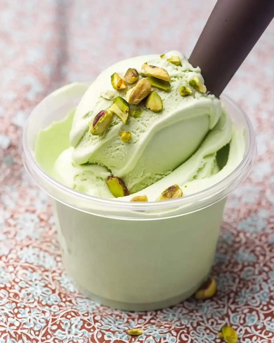 A vibrant scoop of Ninja Creami Pistachio Ice Cream in a clear container, garnished with chopped pistachios, with a dark utensil.