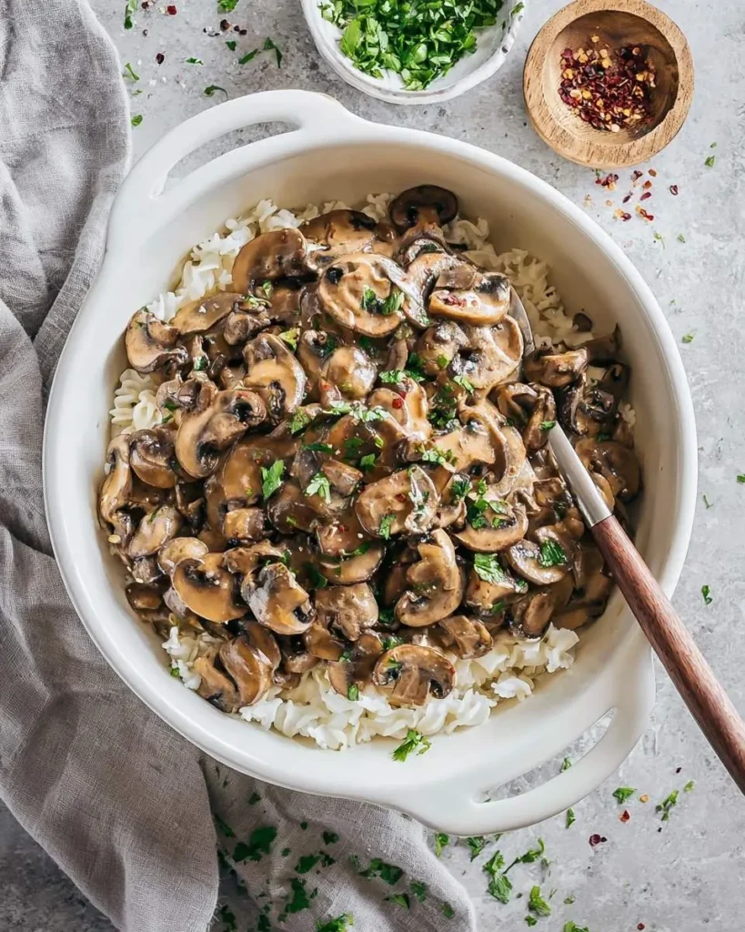 A white casserole dish holds fluffy white pasta topped with creamy Vegan Mushroom Stroganoff, garnished with parsley and red pepper flakes.
