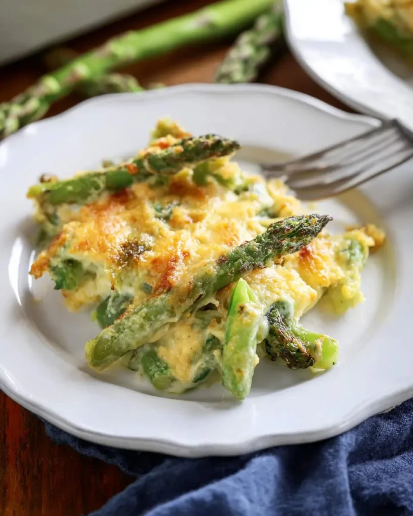 A close-up of a serving of cheesy asparagus casserole with roasted asparagus spears and a golden-brown baked topping on a white plate.