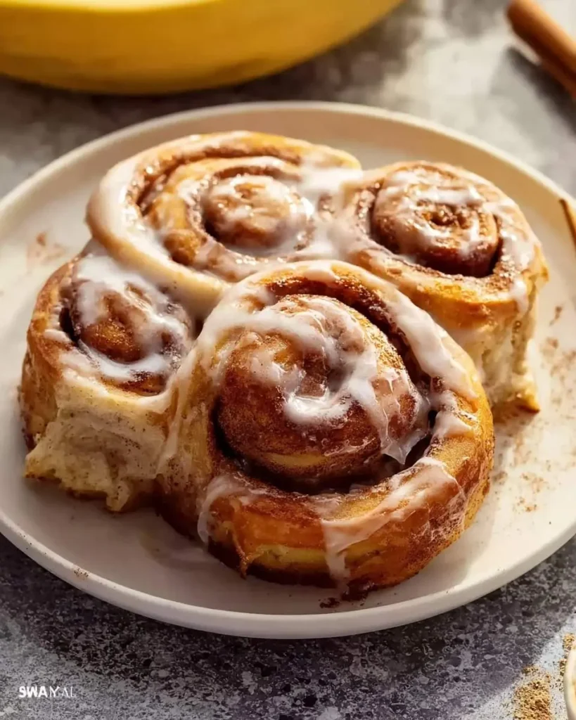Four irresistible Banana Bread Cinnamon Rolls, generously drizzled with white icing, served on a plate with a banana in the background.