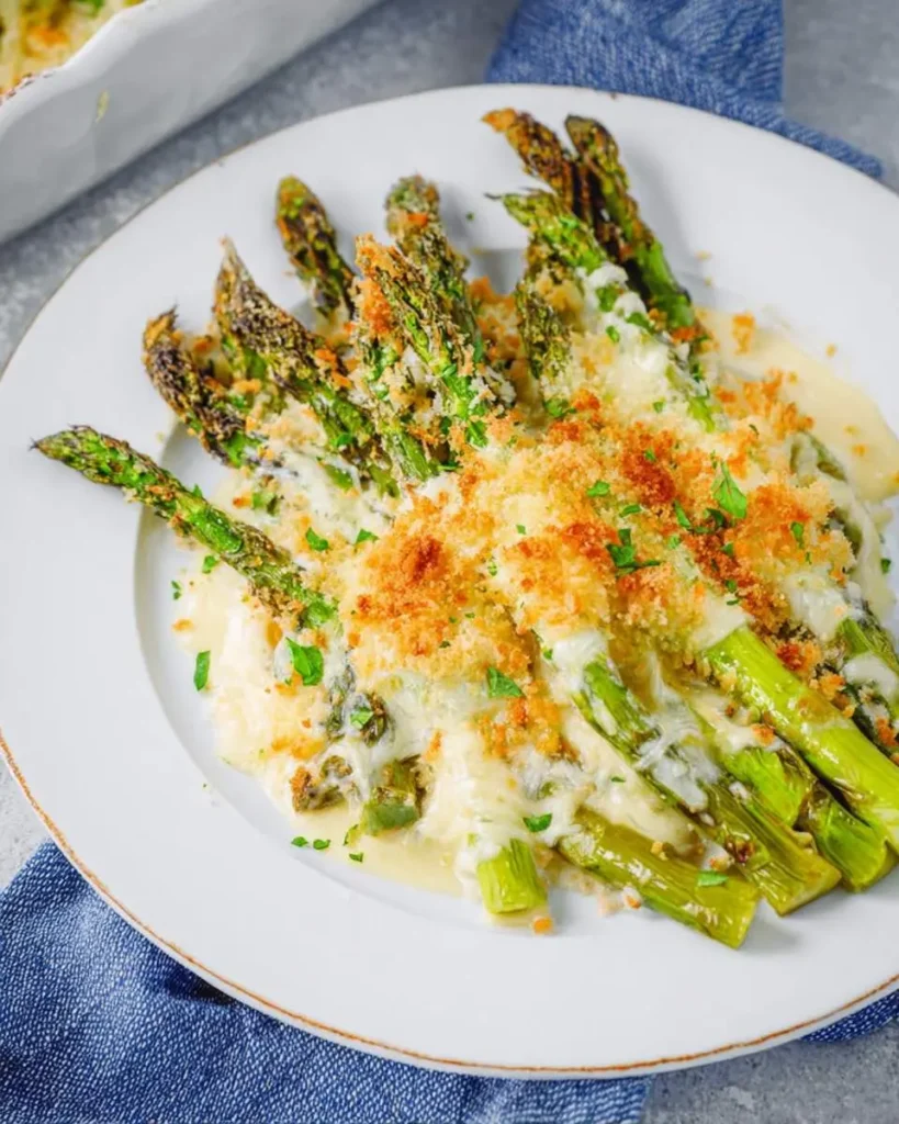 A white plate filled with vibrant green Cheesy Asparagus casserole, topped with melted white cheese, golden breadcrumbs, and fresh parsley.