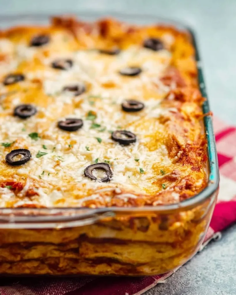 A close-up of a bubbling, cheesy chicken enchilada casserole in a glass baking dish, topped with black olives and fresh herbs.