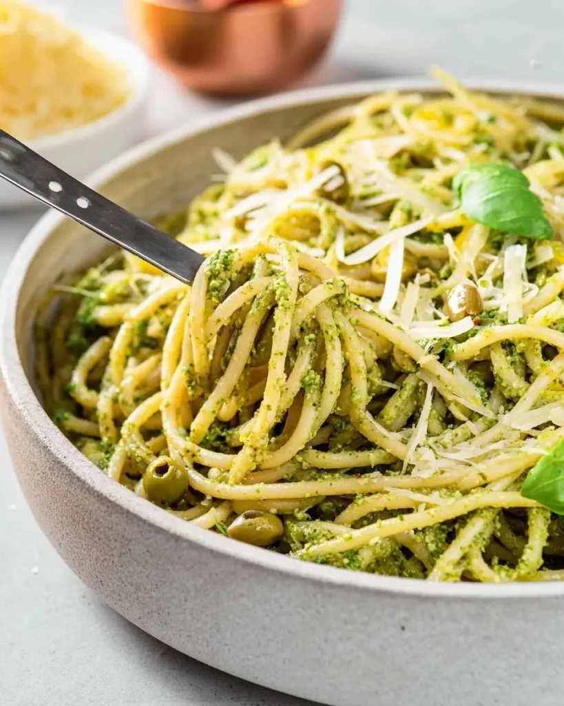 A fork lifts spaghetti coated in vibrant Green Olive Pasta Sauce, topped with grated cheese and basil, from a stone bowl.