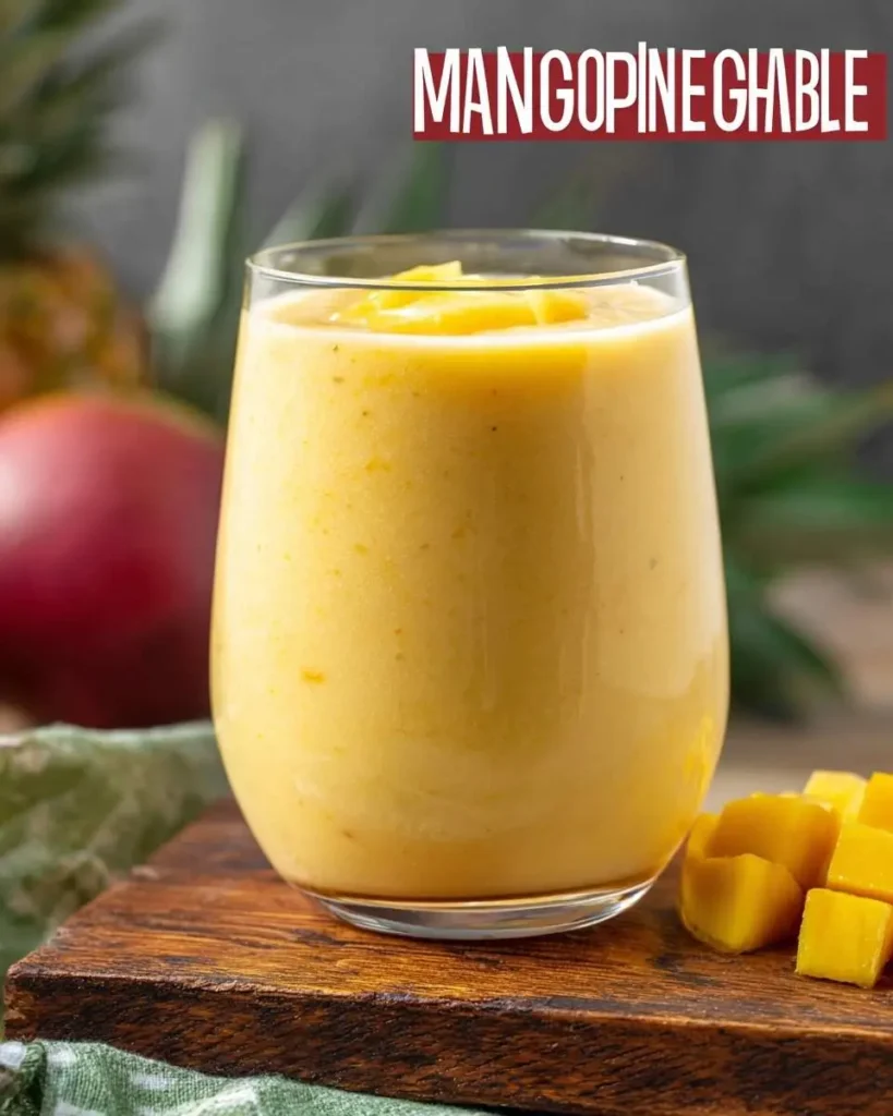 A refreshing Mango Pineapple Smoothie served in a clear glass, garnished with fruit, on a wooden board with fresh mango chunks.