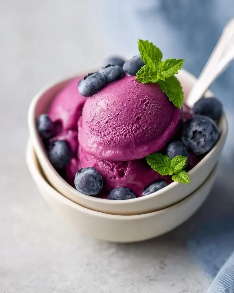 Scoops of vibrant purple Ninja Creami Blueberry Frozen Yogurt garnished with fresh blueberries and mint in a white bowl.
