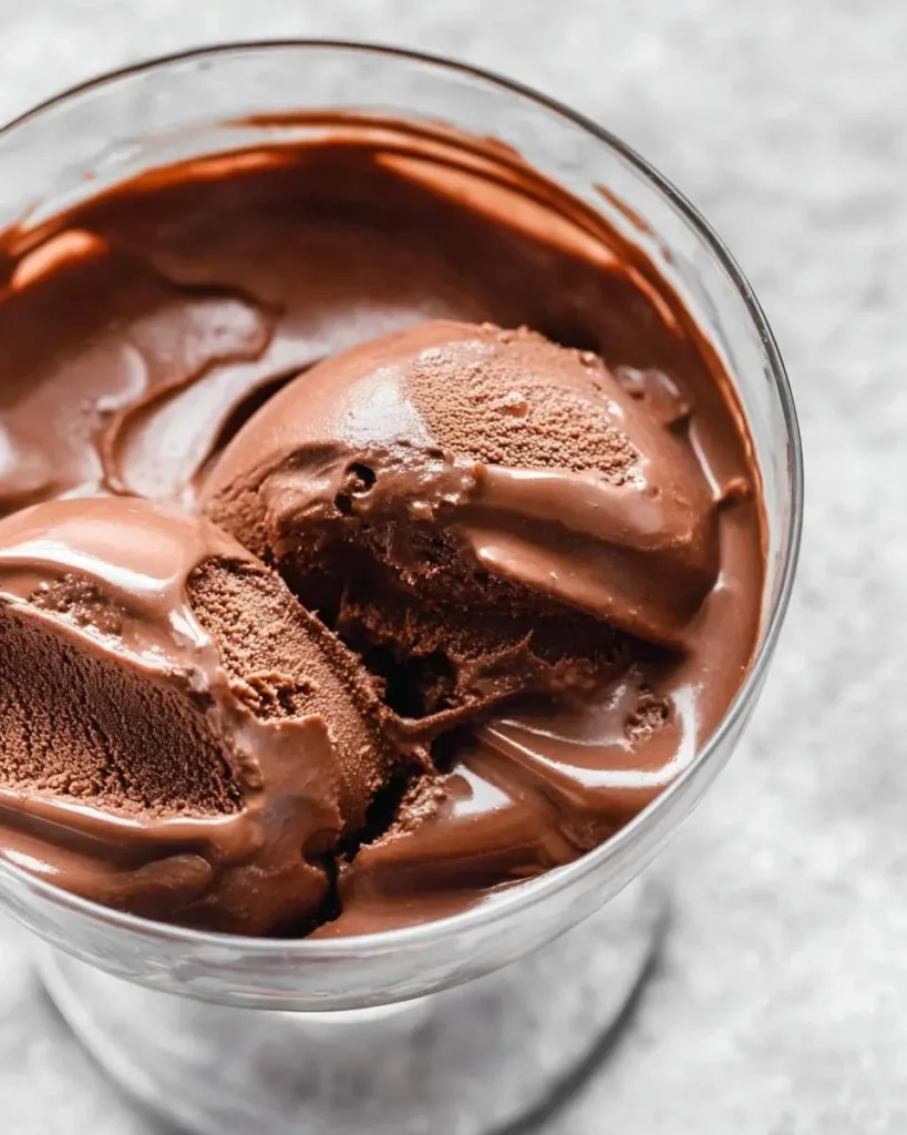 A close-up of two scoops of rich, dark Ninja Creami Chocolate Ice Cream slightly melting in a clear glass dessert bowl.