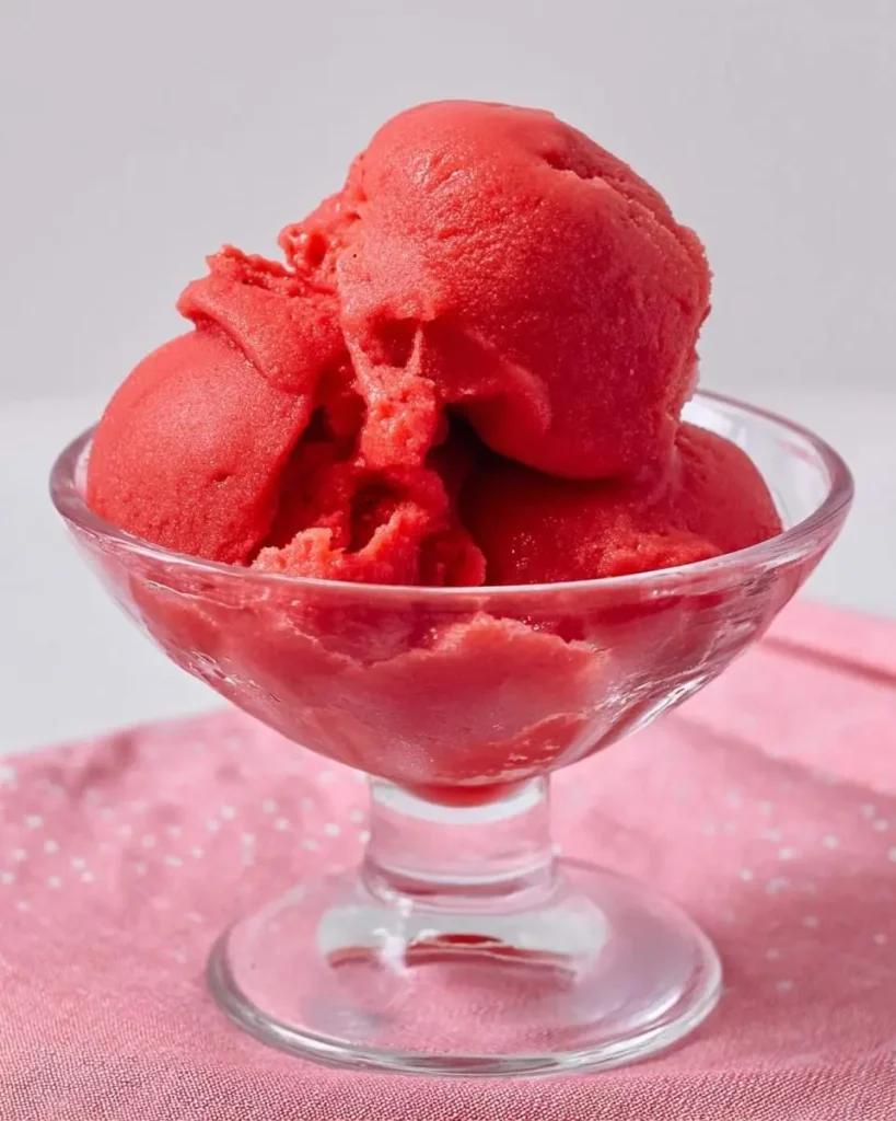 A clear glass dessert bowl filled with vibrant red Ninja Creami Strawberry Sorbet, placed on a pink polka dot napkin.