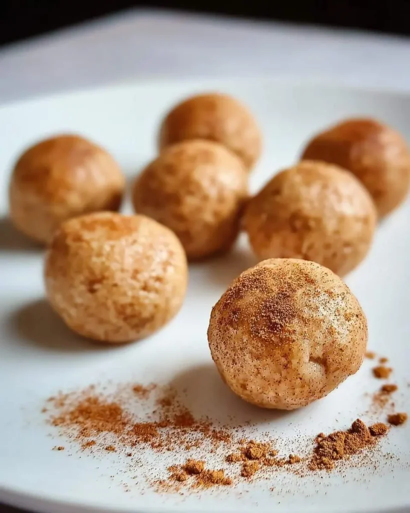 A white plate featuring several golden-brown Snickerdoodle Protein Balls, with one in the foreground dusted with cinnamon.