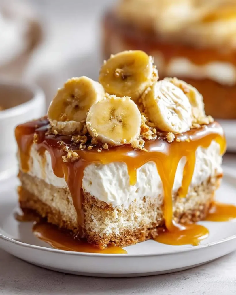 A delectable slice of Bananas Foster Poke Cake, generously topped with fresh banana slices, crunchy bits, and rich caramel sauce dripping down its creamy layers.