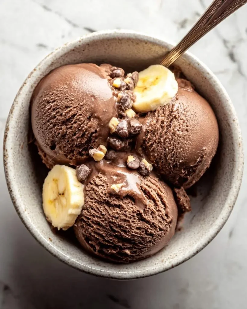 Three scoops of creamy Chocolate Banana Ice Cream topped with fresh banana slices, chocolate chips, and chopped nuts in a ceramic bowl.