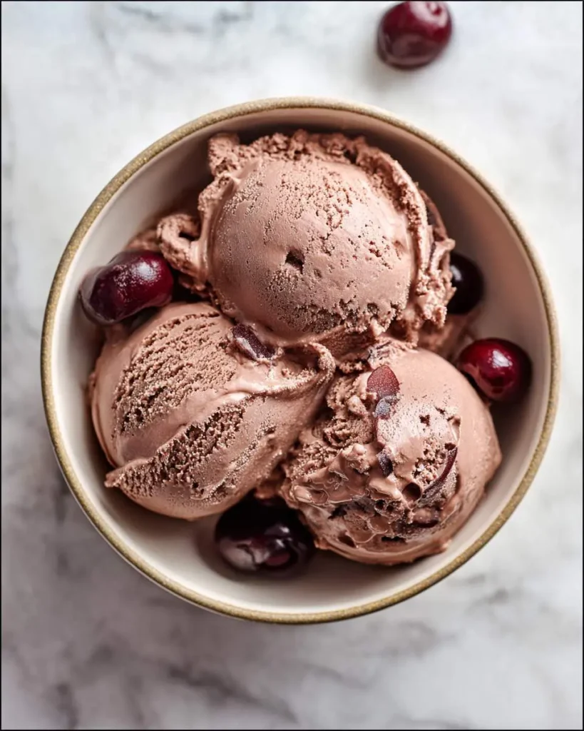 A close-up of a bowl filled with three scoops of rich chocolate cherry ice cream, garnished with fresh red cherries.