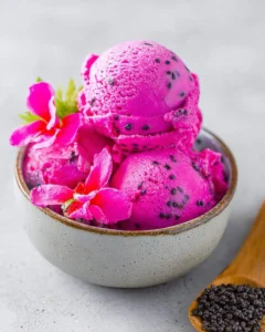 A close-up of vibrant pink Dragon Fruit Ice Cream scoops in a bowl, garnished with fresh pink flowers and black seeds.