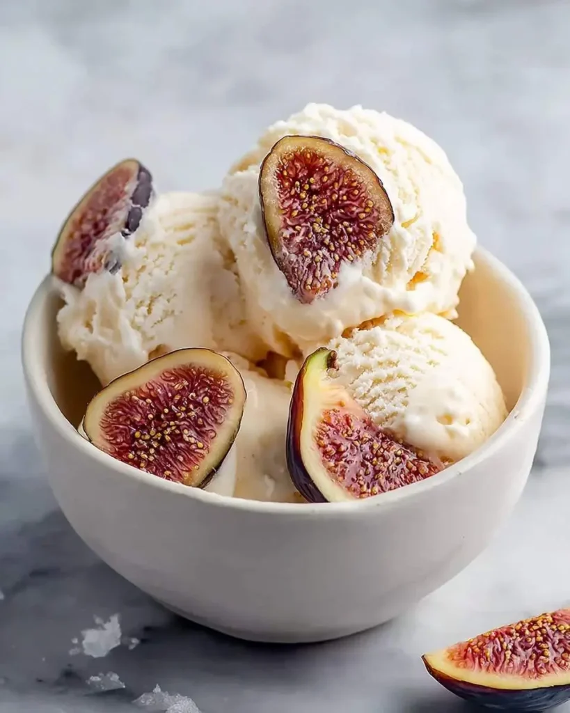 A white bowl filled with creamy scoops of Fig Ricotta Ice Cream garnished with fresh fig halves on a light marble surface.